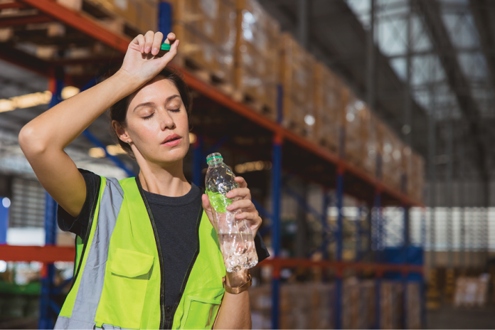 A warehouse worker in a reflective vest wipes her forehead while holding a water bottle.