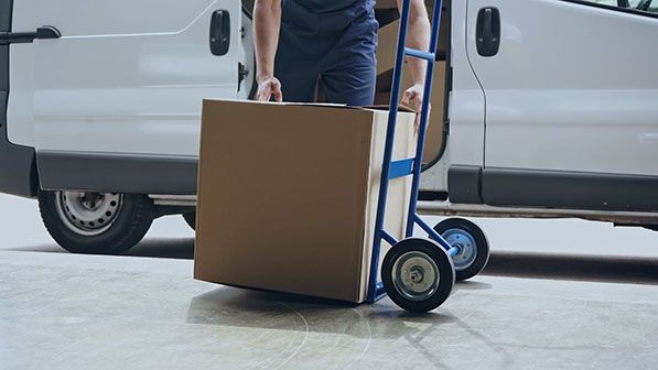 Person moving a large cardboard box on a blue hand truck near a white delivery van.