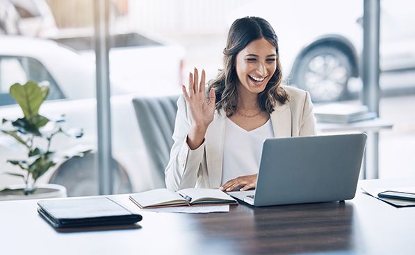 A smiling woman waves toward her laptop camera while seated at a desk.