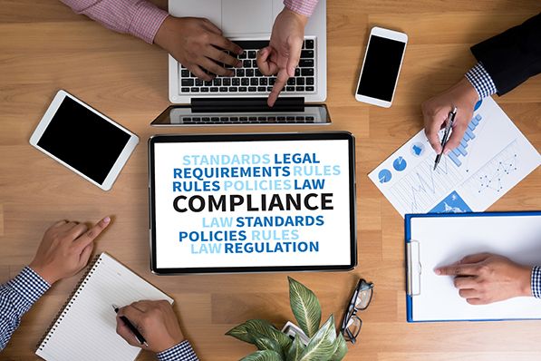Overhead view of coworkers around a desk with a tablet showing a compliance-related word cloud.