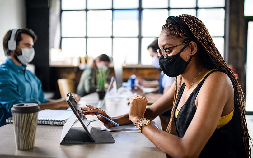 Masked coworkers sit at a shared table, with a woman in the foreground using a tablet.