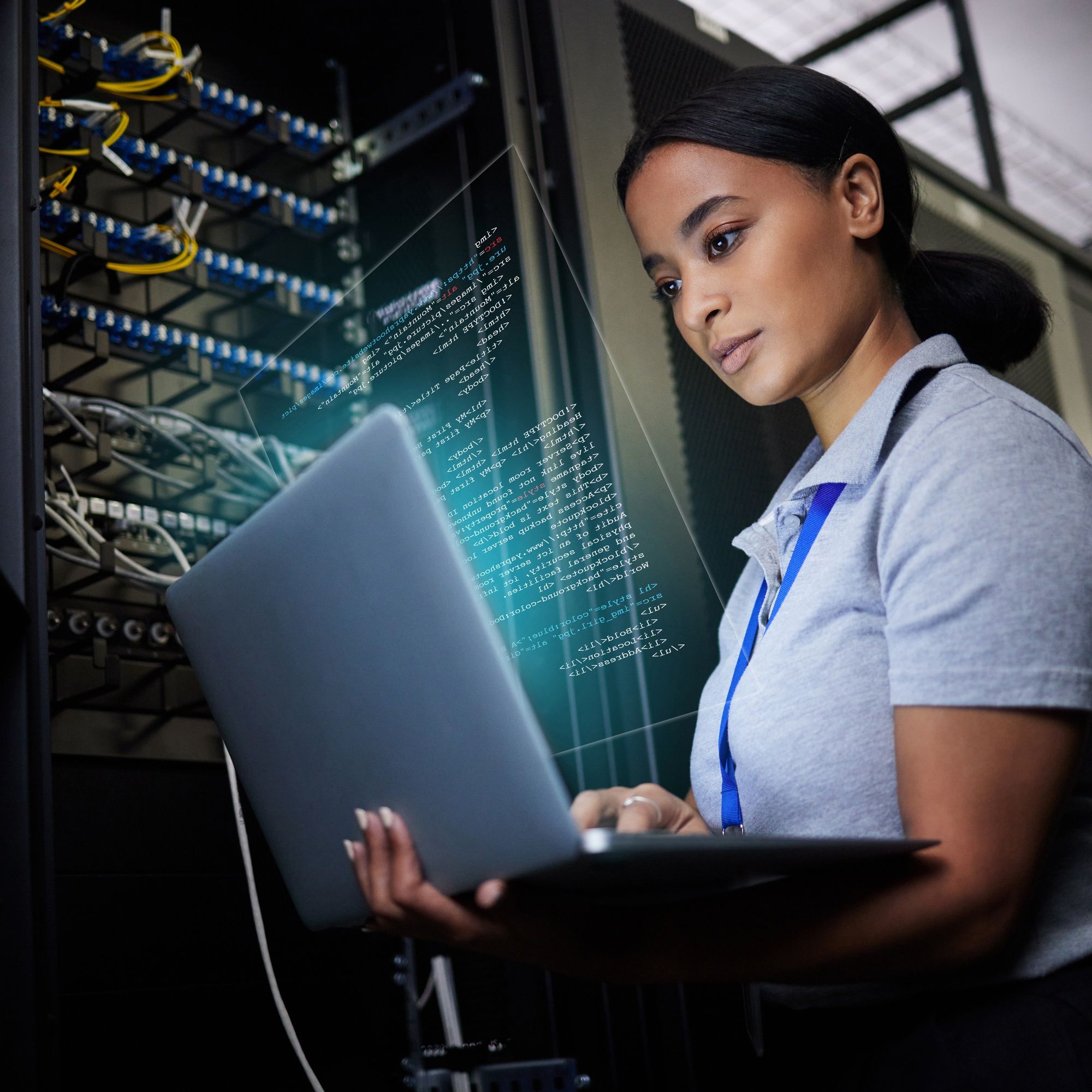 A woman uses a laptop beside racks of network equipment.
