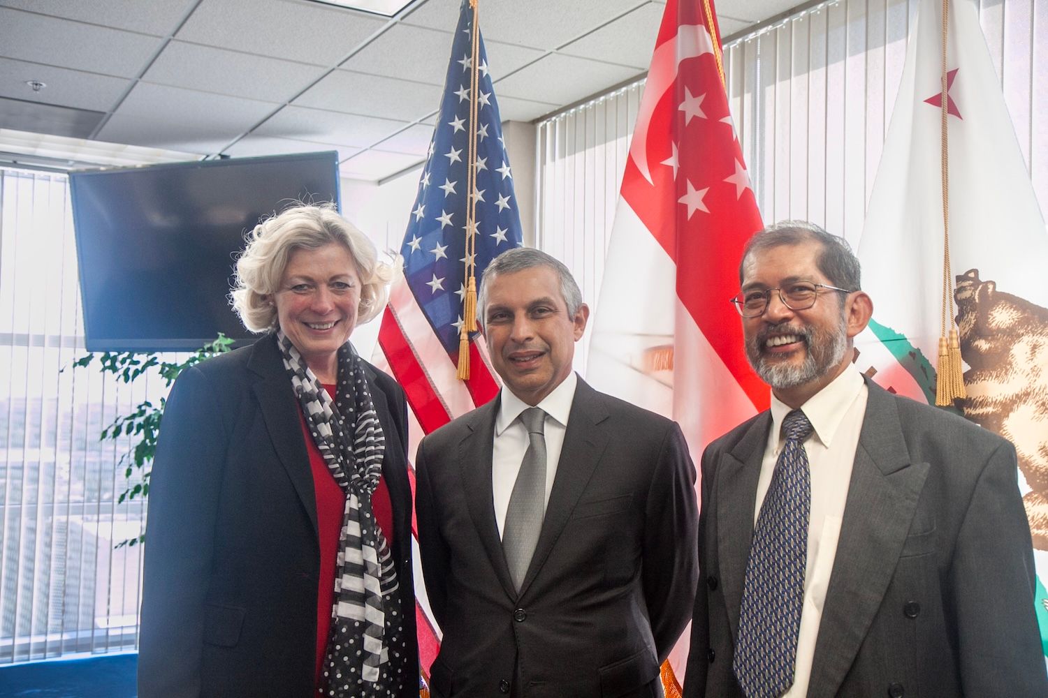 Three people in business attire pose before United States, Singapore and California flags.