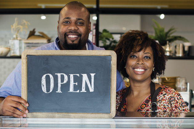 Two smiling shop workers hold an OPEN chalkboard behind a counter.