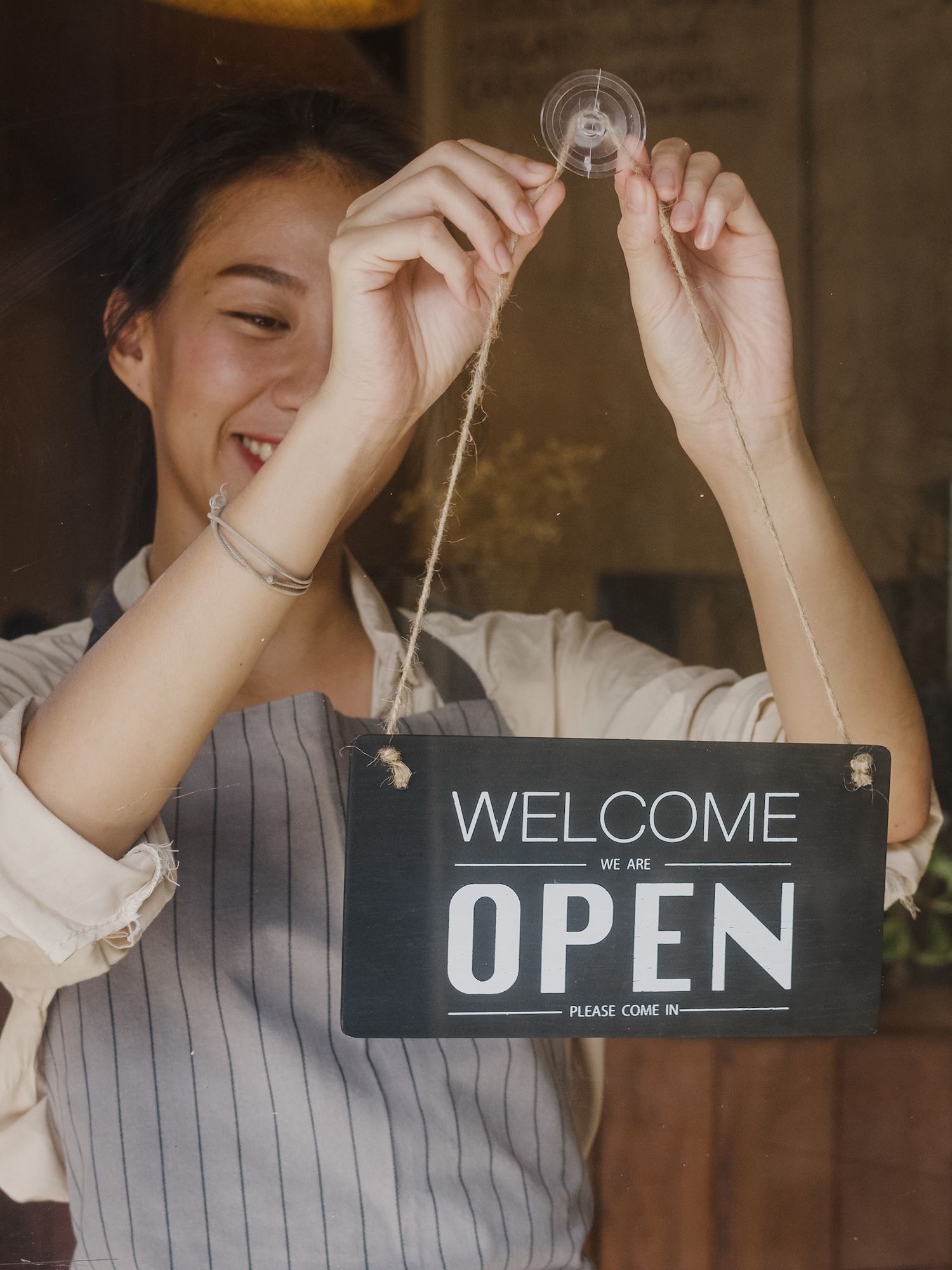 A smiling worker in an apron hangs a Welcome Open sign on a glass door.