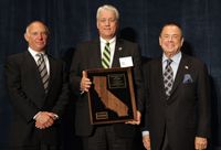 Three men in suits pose together, with the center man holding a framed California award.