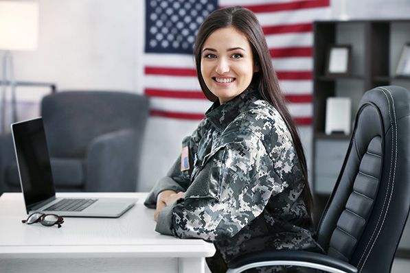 A woman in camouflage uniform sits at an office desk with a United States flag behind her.