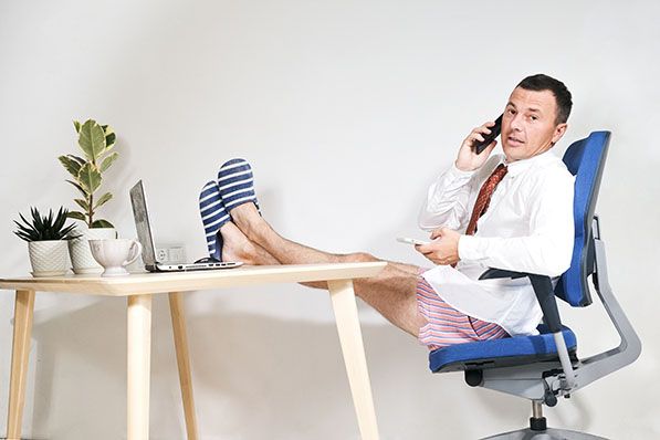 A man in a shirt, tie, shorts and slippers talks on a phone with his feet on a desk.
