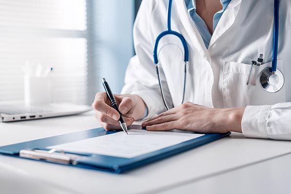 A clinician in a white coat and stethoscope writes on a clipboard.