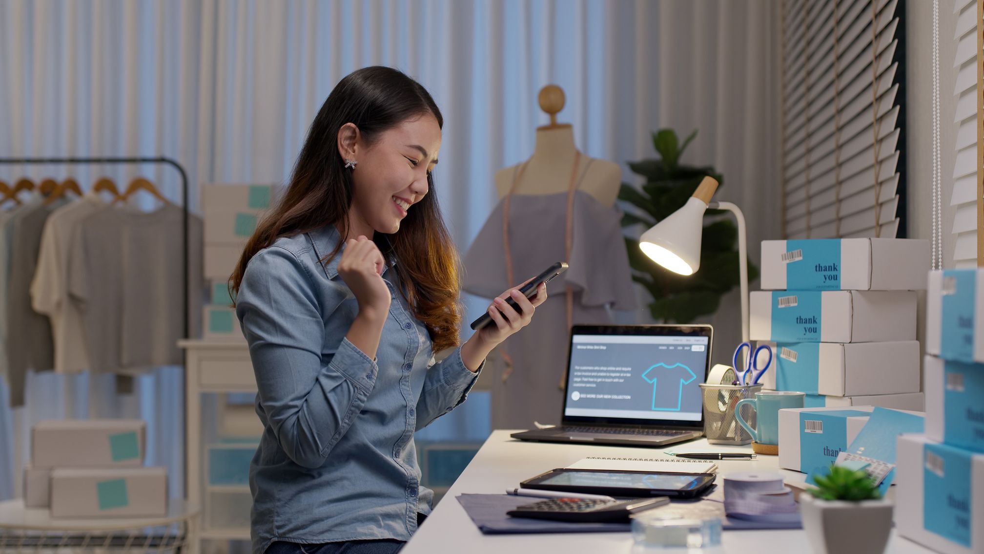 Happy woman celebrating while looking at her phone in a home office with clothing and shipping boxes.
