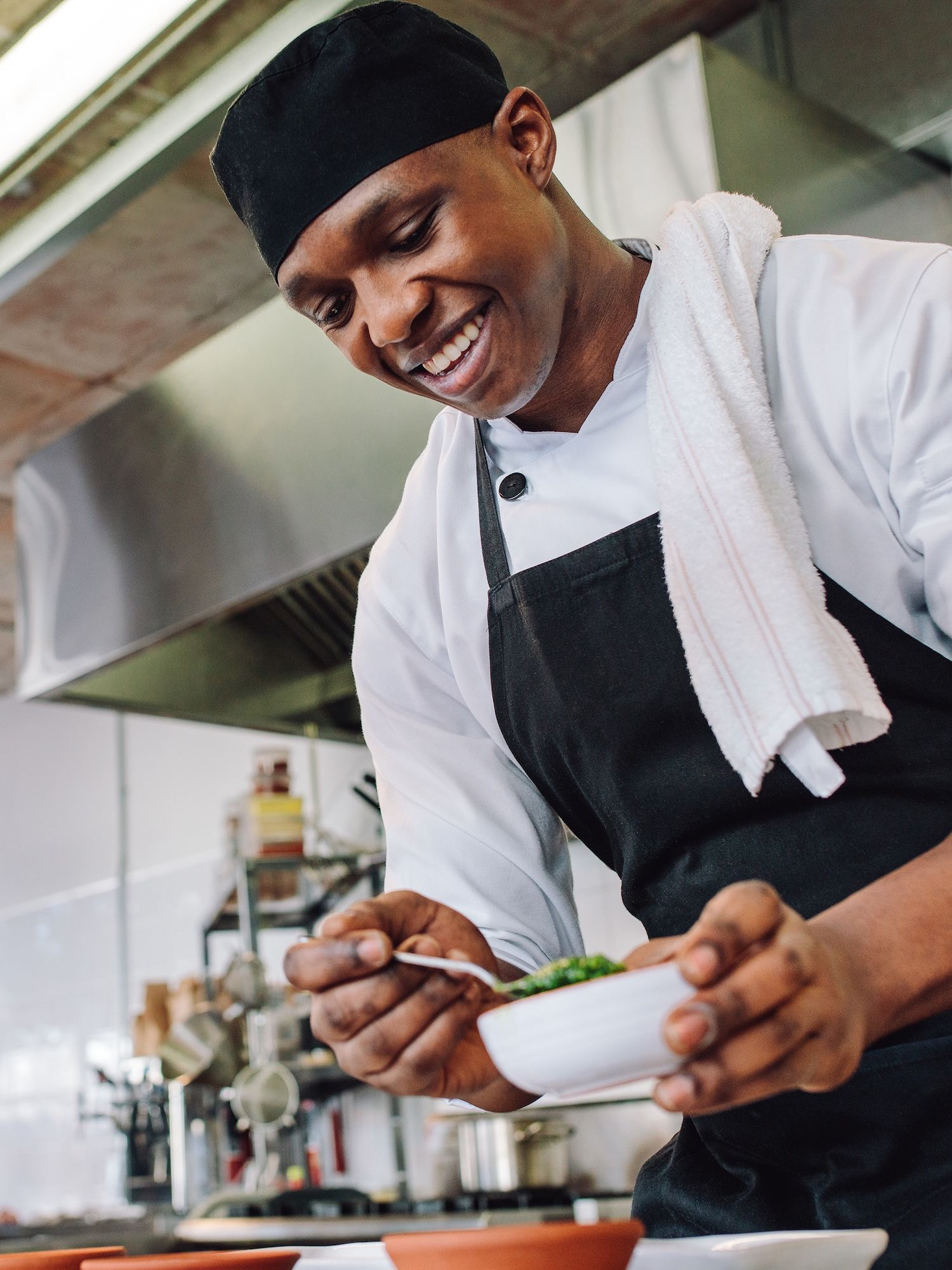 A smiling chef in a white jacket and black apron prepares food in a commercial kitchen.
