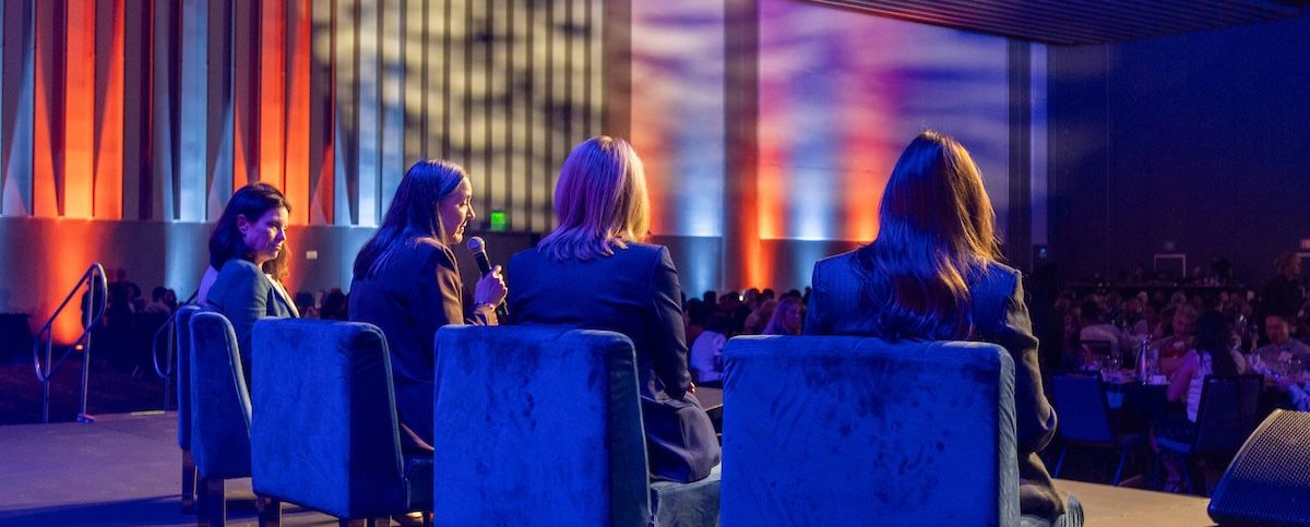 Panelists seated onstage face a large audience in a hall lit blue and orange.