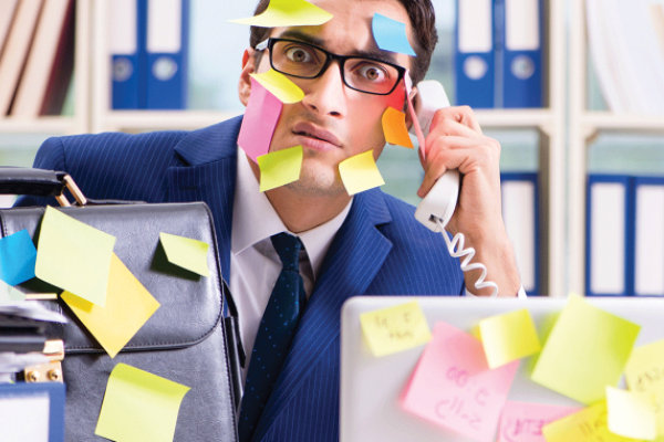 A man in glasses holds a telephone amid sticky notes covering his face, clothing and desk equipment.
