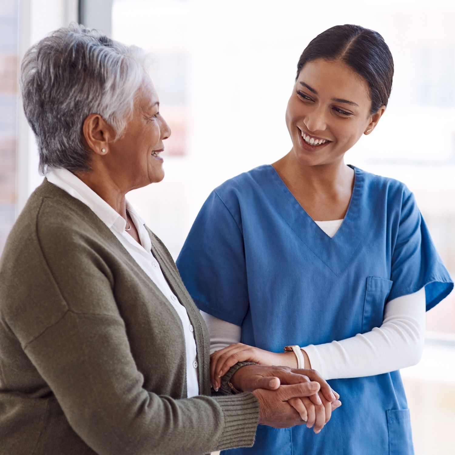A healthcare worker in blue scrubs holds the hands of a seated older adult.