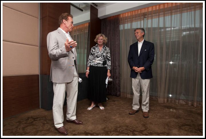 Three people stand in a meeting room while a man in a light suit gestures as he speaks.