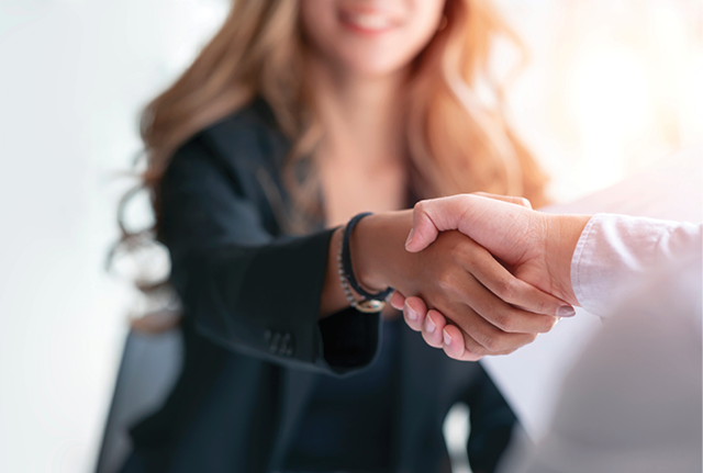 Close-up of a handshake with a smiling woman blurred in the background.