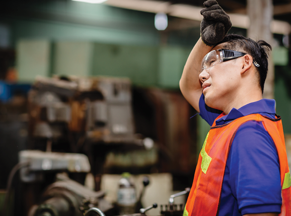 A worker in safety glasses and an orange vest wipes their forehead with a gloved hand.