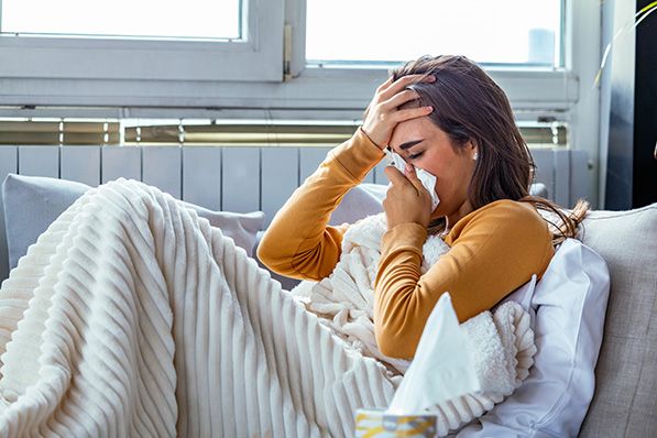 A woman sits under a blanket on a couch, holding a tissue to her nose and a hand to her forehead.