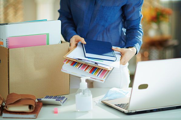 A person packs notebooks and folders beside a cardboard box and laptop on an office desk.