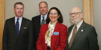Four people in business attire pose together indoors, including a woman in a red jacket.