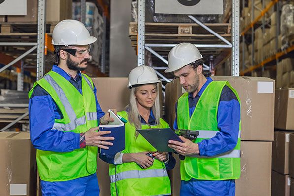 Three warehouse workers in hard hats and reflective vests review a clipboard and handheld device.
