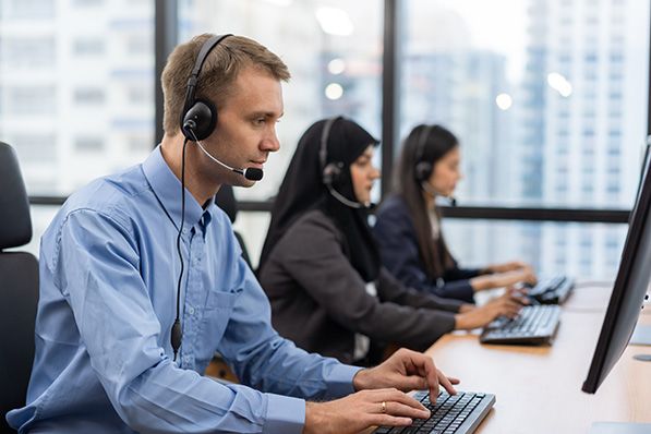 Three office workers wearing headsets sit at computers in a row.