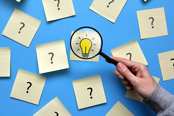 A hand holds a magnifying glass over a sticky note with a lightbulb, surrounded by sticky notes with question marks on a blue surface.