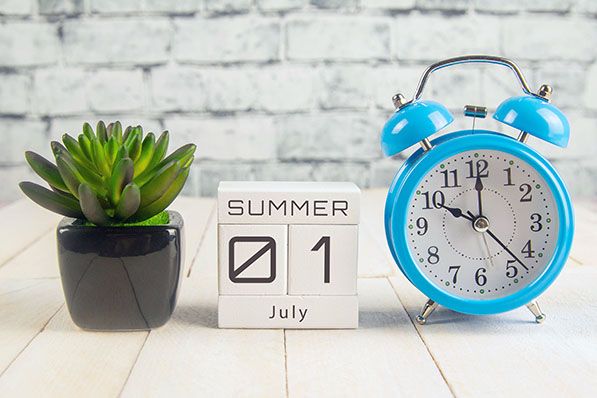 A blue alarm clock and potted succulent flank a block calendar set to July 1.