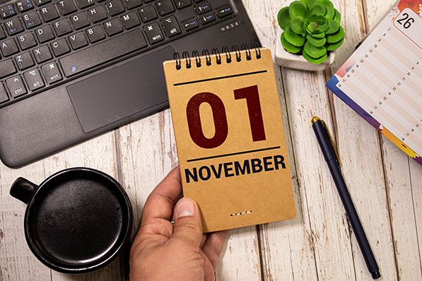 A hand holds a desk calendar showing November 01 beside a laptop, coffee cup and stationery.