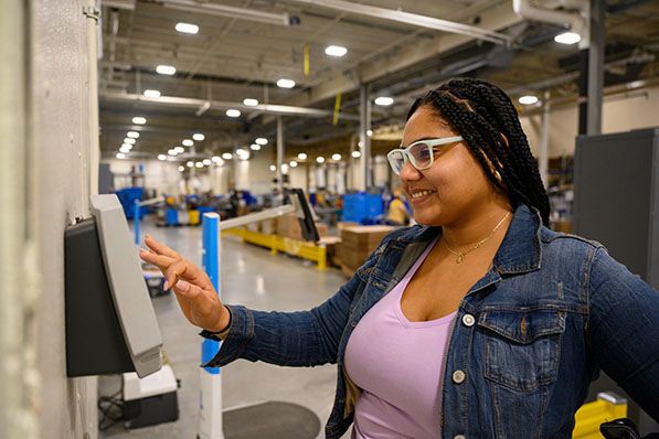 A smiling worker taps a wall-mounted touchscreen in a warehouse.