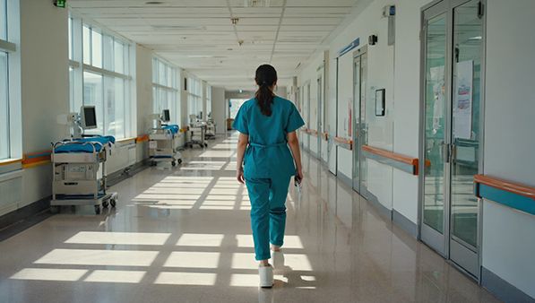 A healthcare worker in teal scrubs walks down a bright hospital corridor.