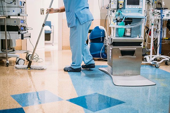 A worker in blue scrubs mops a hospital floor beside medical equipment.