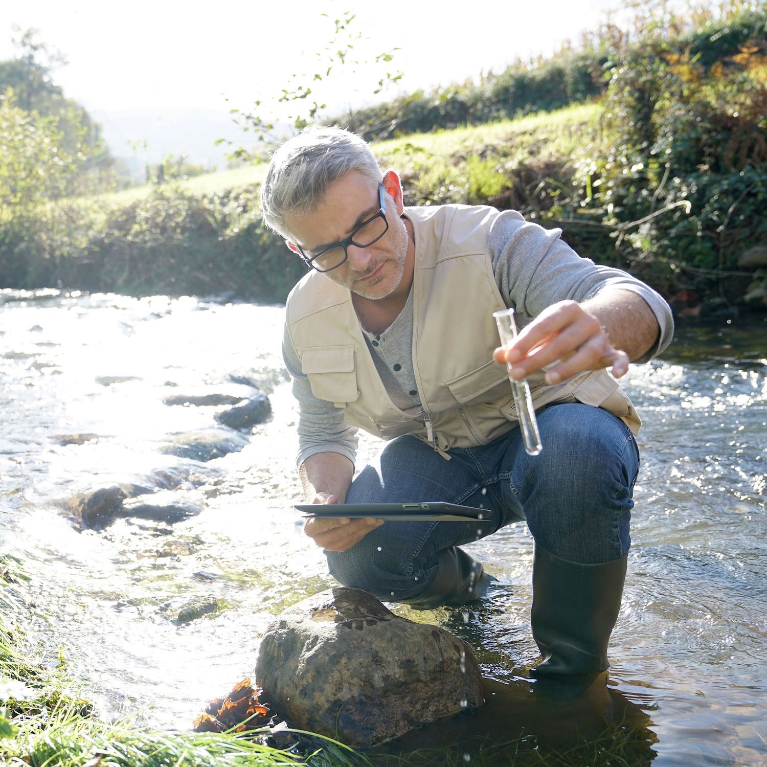 A person kneels beside a stream, holding a sample tube and recording information on a tablet.
