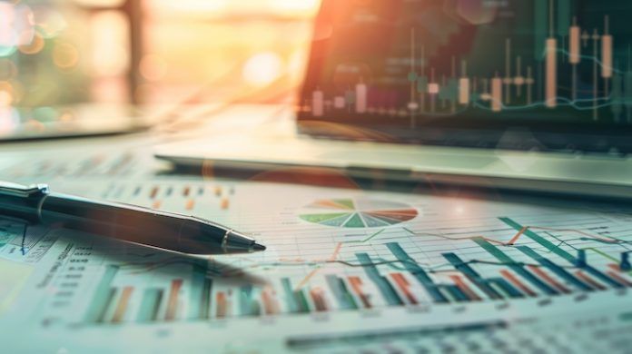 Financial charts and a pen lie on a desk in front of a laptop displaying market data.