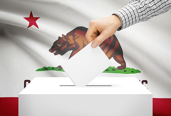 A hand inserts a ballot into a white box in front of a California Republic flag.