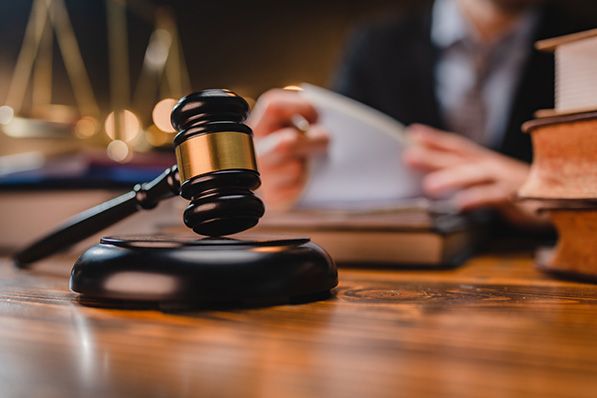 A judge's gavel rests on a desk with a blurred person reviewing documents behind it.