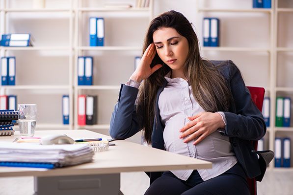 A pregnant woman sits at an office desk with one hand on her abdomen and the other at her temple.