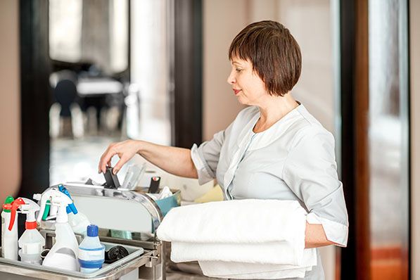 A hotel housekeeper arranges supplies on a cart stacked with towels and cleaning products.
