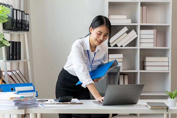 A woman holding a blue folder leans over a laptop at an office desk.