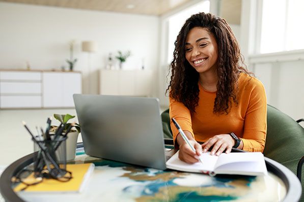 A smiling woman uses a laptop and writes in a notebook at a table.