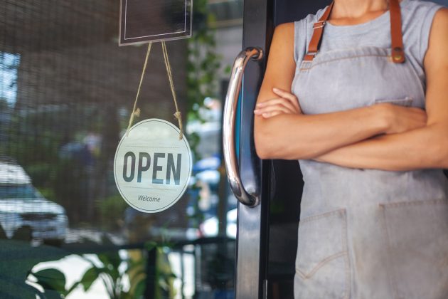 A person wearing an apron stands with folded arms beside a shop door displaying an OPEN sign.