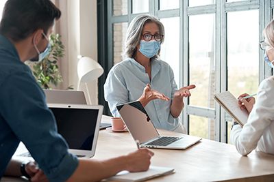 Masked coworkers discuss around a table with laptops and notes in a bright office.