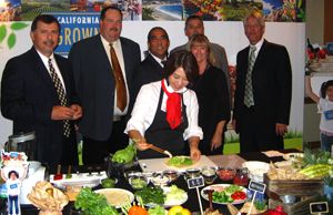 A cook prepares food at a display table surrounded by produce and suited attendees.