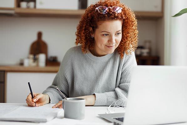 A woman with curly red hair writes notes while looking at a laptop at home.