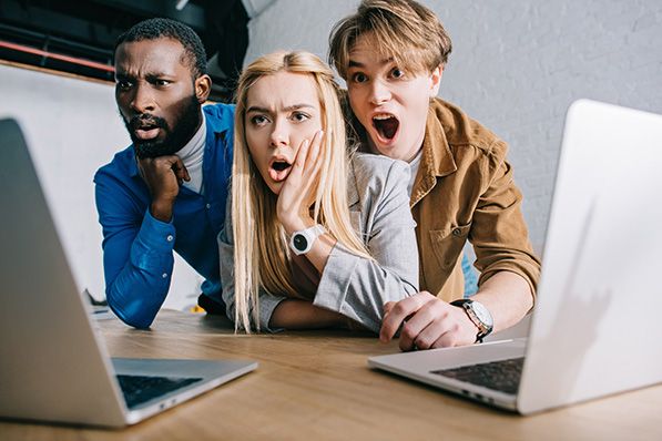 Three people lean toward a laptop with widened eyes and open mouths.