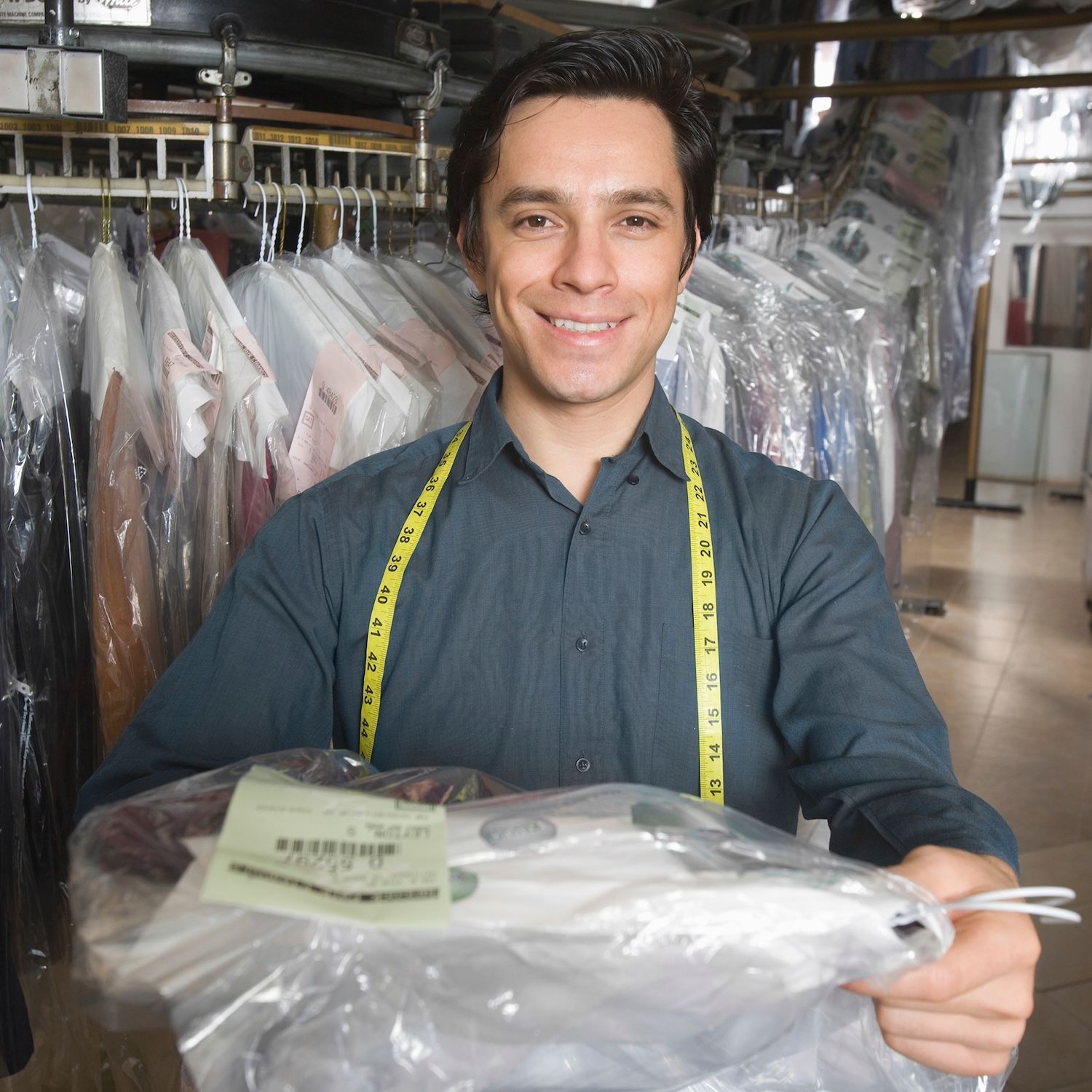 A smiling dry-cleaning worker with a measuring tape around his neck holds a packaged garment.