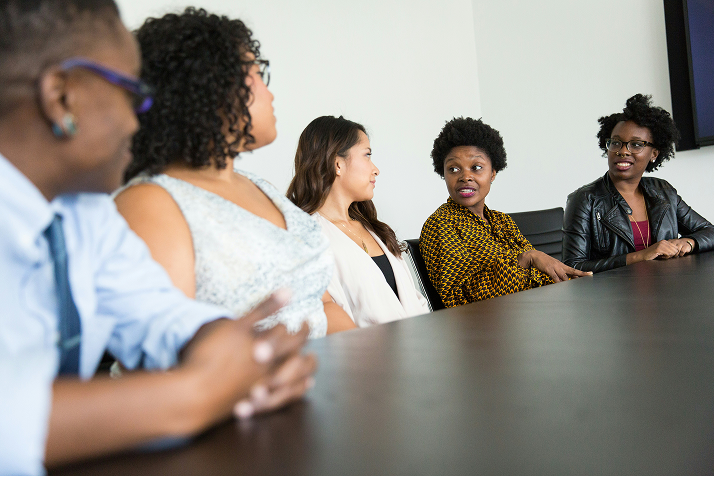 Five people sit together at a conference table, talking during an office meeting.