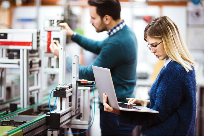 Two workers inspect industrial equipment, with one using a laptop.