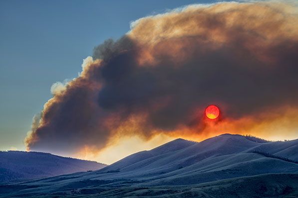 A thick smoke plume stretches across the sky above hills and a red sun.
