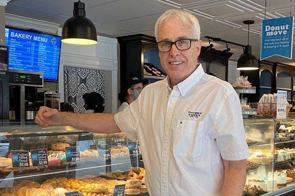 Kirk Rossberg stands beside a pastry display counter in a white short-sleeved shirt.
